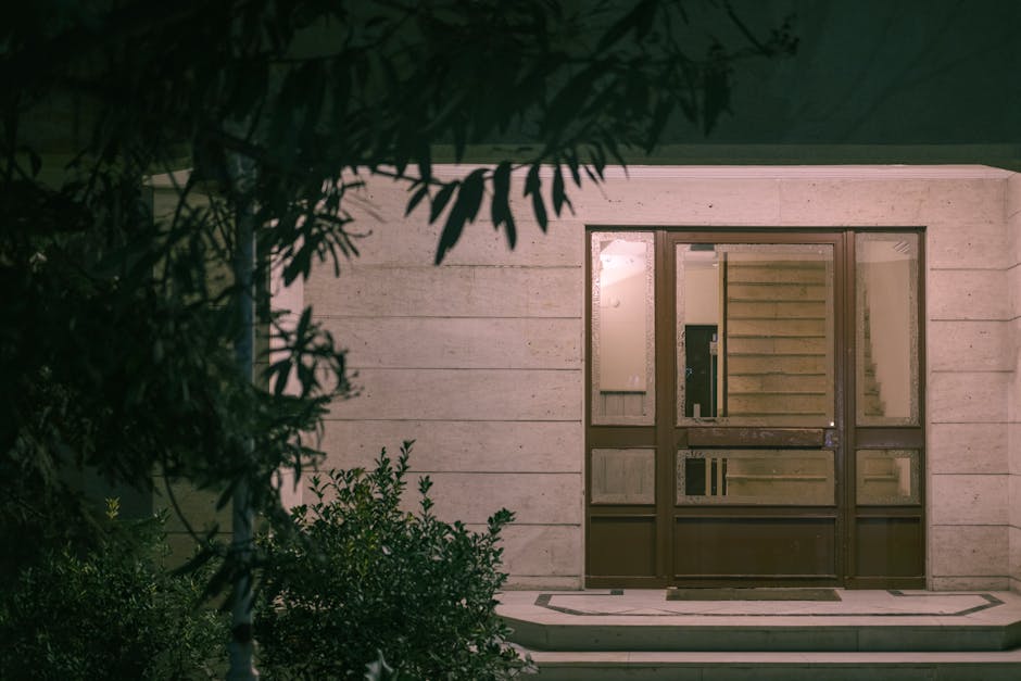 Une vue nocturne apaisante de l'entrée d'un bâtiment moderne entouré de végétation, créant une atmosphère chaleureuse et accueillante.